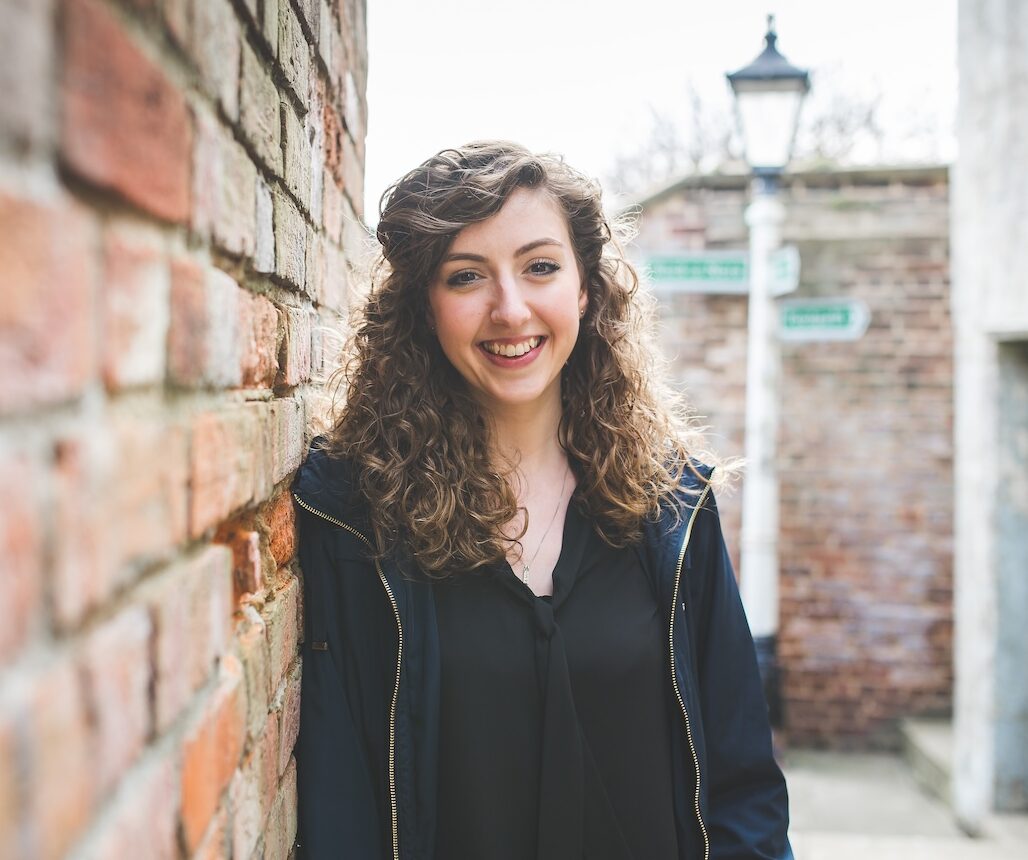 Smiling woman with curly hair leaning against a brick wall outdoors