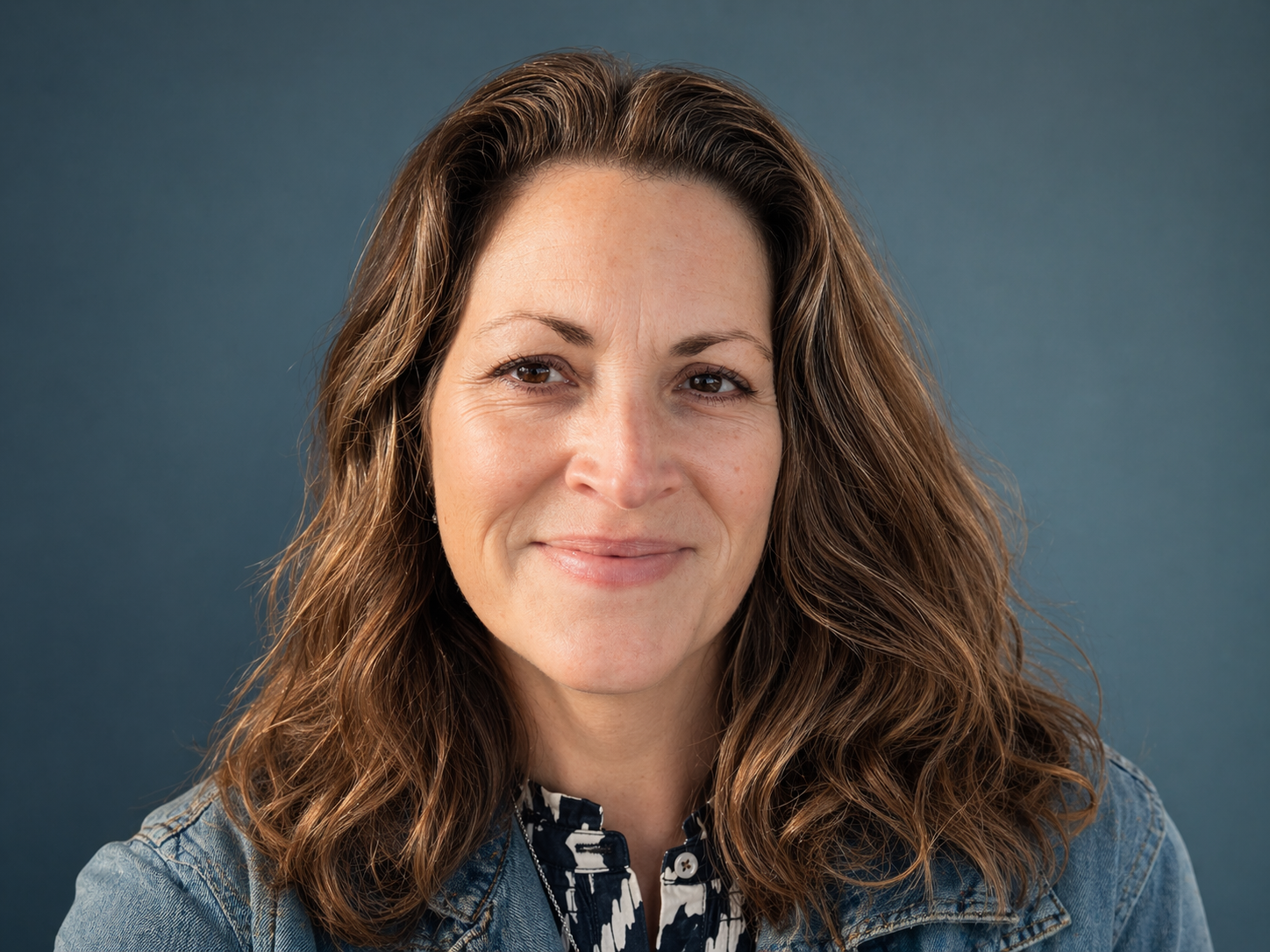 Portrait of a woman with wavy brown hair wearing a denim jacket and patterned blouse