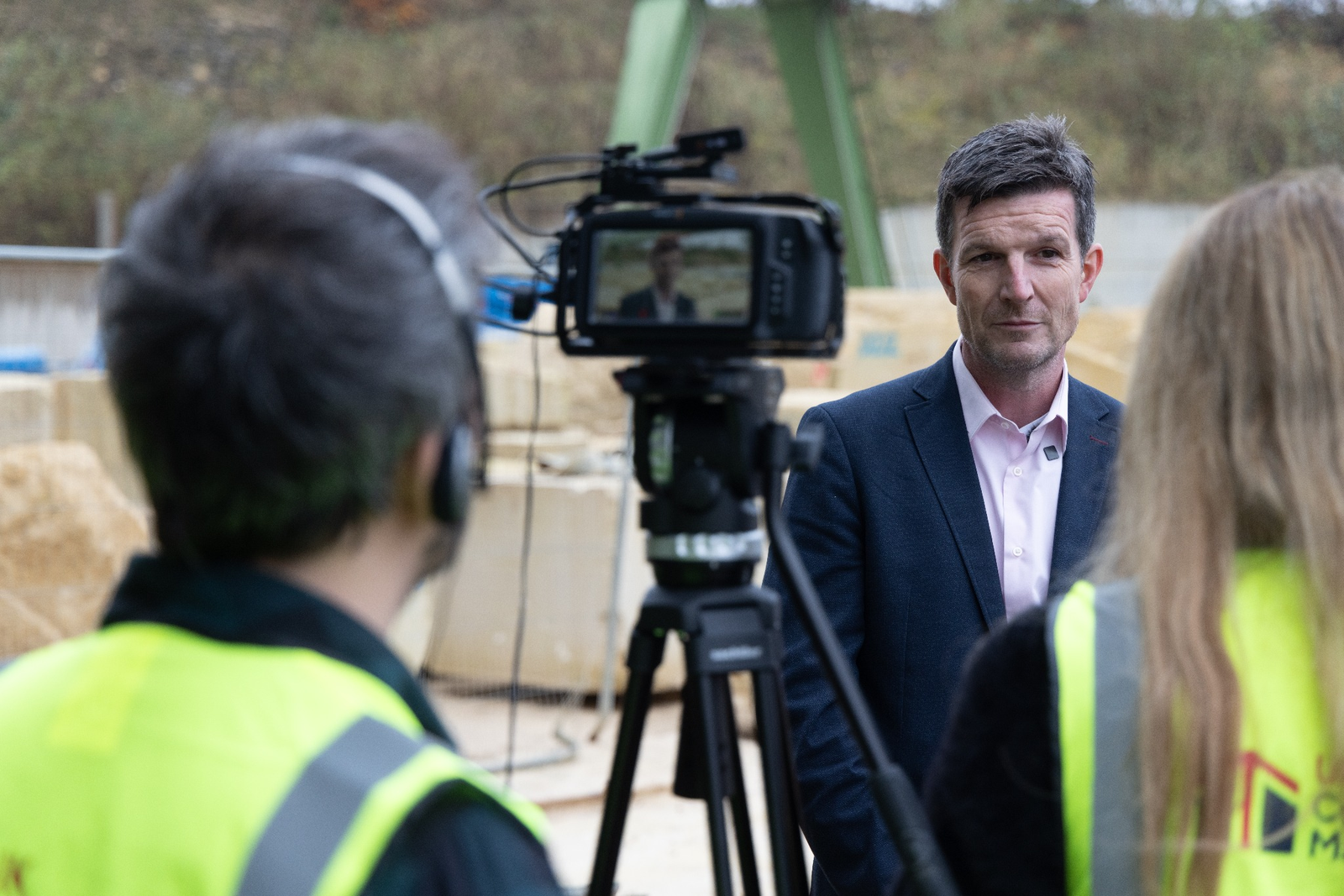Man in suit being filmed by two people wearing high-visibility vests on a construction site