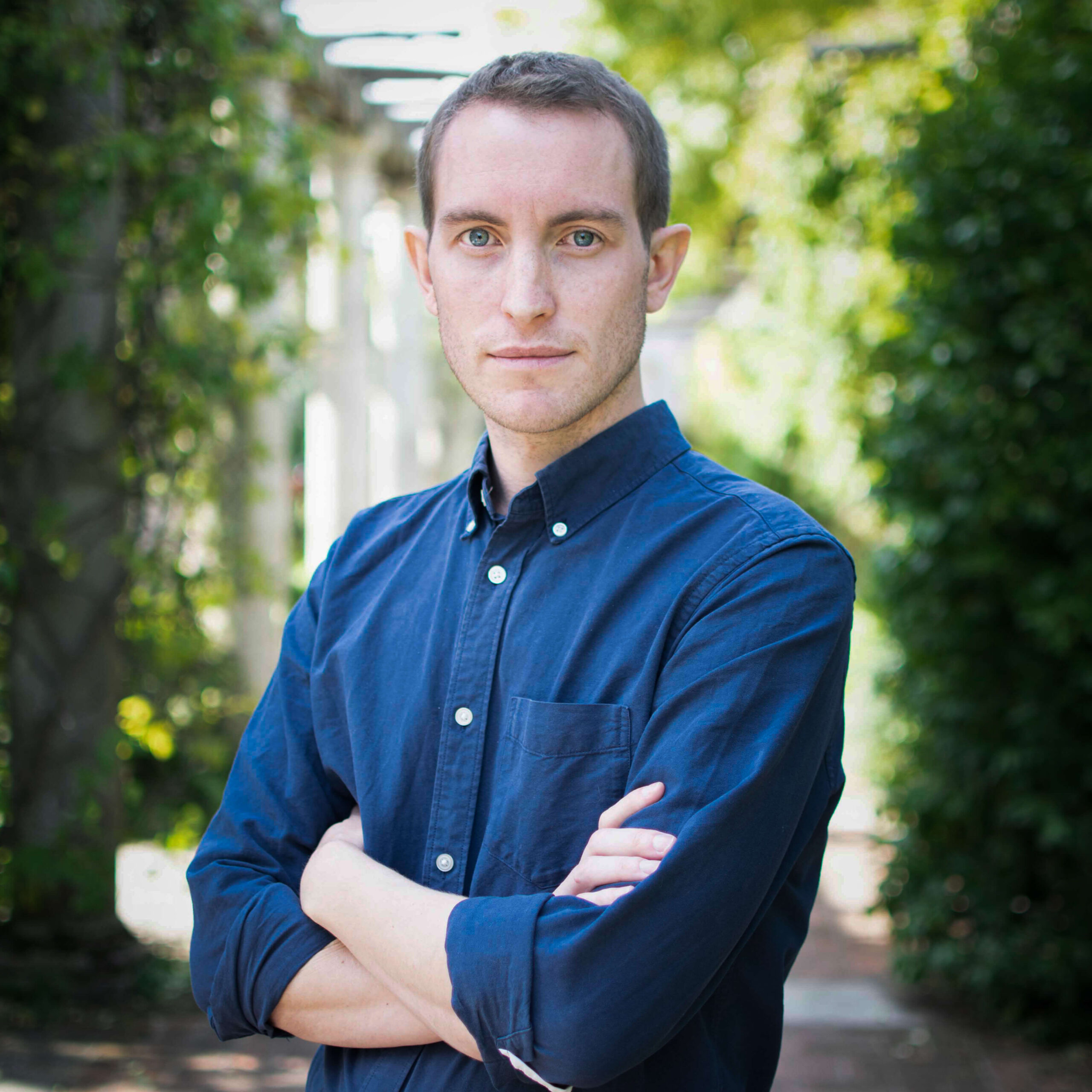 Man with short hair wearing a blue button-up shirt standing outdoors with arms crossed