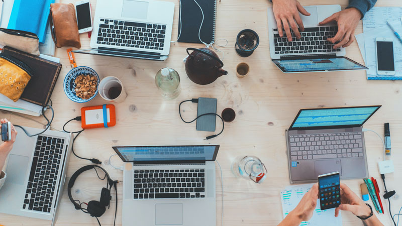 Overhead view of a wooden table with multiple laptops, smartphones, notebooks, and coffee cups during a team work session